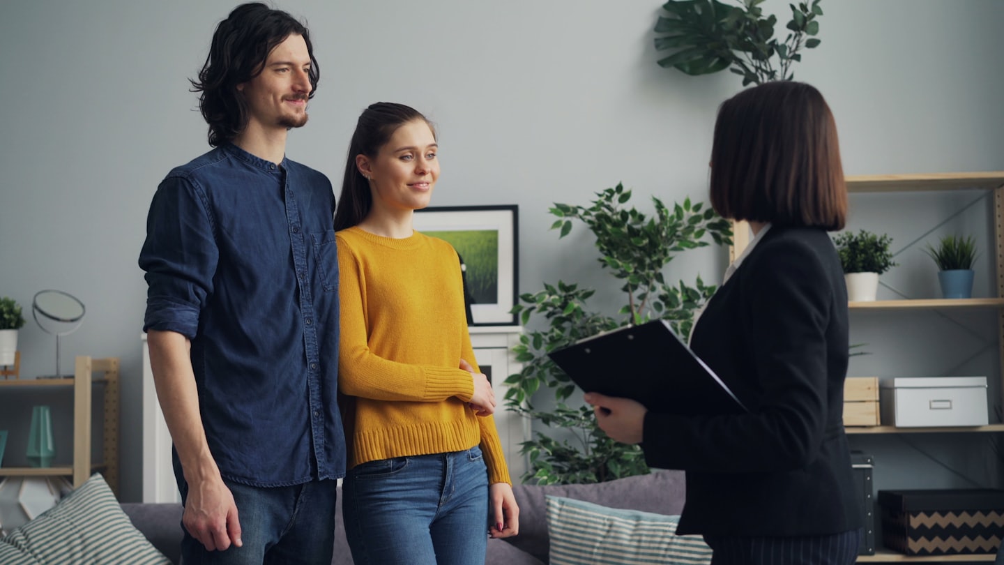 Couple listens to agent holding a clipboard in a modern living room.